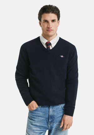Navy v-hals trui gemaakt van zacht materiaal, met geribbelde boorden en zoom, gedragen over een witte shirt en stropdas, gecombineerd met lichtblauwe jeans.