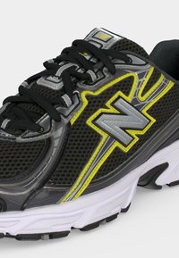 Μαύρο και γκρι παπούτσι τρεξίματος New Balance με κίτρινες λεπτομέρειες, διάτρητο επάνω μέρος, μαύρα κορδόνια και λευκή μαξιλαρωτή σόλα, εμφανιζόμενο από πλάγια εμπρόσθια γωνία.