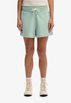 Parte inferiore del corpo di una persona che indossa pantaloncini verdi chiaro con coulisse, calzini bianchi e scarpe da ginnastica bianche alte su uno sfondo uniforme.