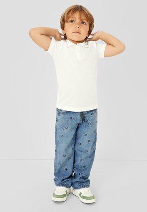 Młode dziecko o jasnobrązowych włosach stoi z rękami za głową, ubrane w białą koszulkę polo, niebieskie jeansy z wzorem oraz białe trampki z zielonymi akcentami.