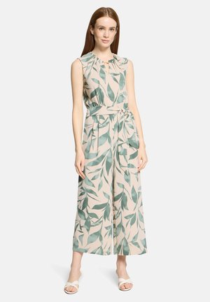 Žena s dlhými červenými vlasmi, oblečená v béžovom overale bez rukávov so vzorom zelených listov, s opaskom v páse a bielymi sandálmi s otvorenou špičkou, stojaca na bielom pozadí.