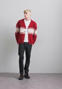 Bărbat tânăr în picioare, îmbrăcat cu un cardigan în dungi roșii și bej, un tricou alb, pantaloni negri și pantofi maro închis, pe un fundal plain gri.