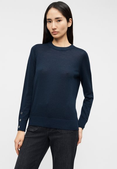 Μπλε ναυτικό πουλόβερ σε απαλό πλεκτό με στρογγυλή λαιμόκοψη, μακριά μανίκια και διακοσμητικά κουμπιά στις μανσέτες. Συνδυασμένο με σκούρο τζιν παντελόνι.