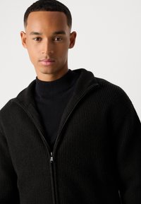 Jeune homme portant un pull zippé noir à côtes sur un col roulé noir, faisant face à l'avant avec une expression neutre sur un fond uni.