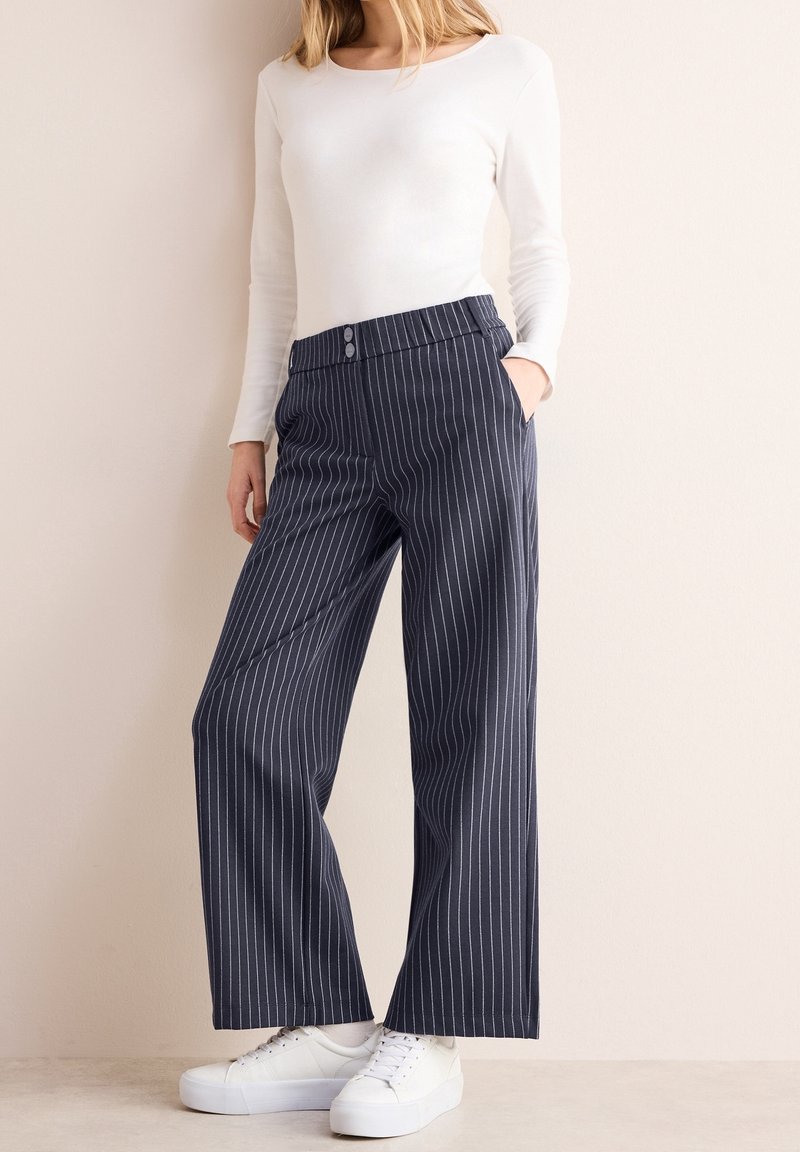 Femme portant un haut blanc à manches longues, un pantalon large à rayures bleu marine avec des boutons à l'avant, et des baskets blanches, debout contre un mur beige.