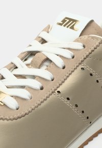 Μπέζ και μεταλλικό χρυσό αθλητικό παπούτσι με λευκά κορδόνια, διάτρητες λεπτομέρειες στα πλαϊνά και λαστιχένια σόλα. Χρυσό λογότυπο στη γλώσσα.