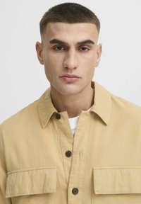Veste en tissu texturé de couleur khaki clair, dotée d'un col, d'une fermeture à boutons et de deux poches avant avec des boutons foncés.