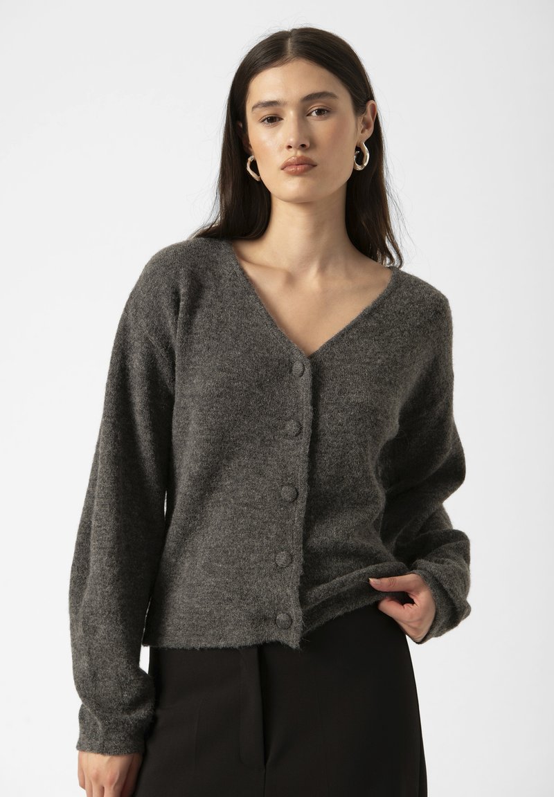 Una giovane donna con lunghi capelli scuri indossa un cardigan grigio con bottoni, in piedi contro uno sfondo bianco puro.