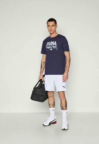 T-shirt marine avec l'imprimé "PUMA SPORTS 1948", shorts blancs, sac de sport noir et baskets blanches avec des accents rouges. Fond uni.