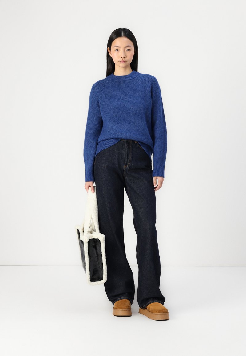 Paarse gebreide trui, donkere denim wijdlopende broek, bruine suède loafers en een zwart-wit schapenvacht tote tas.