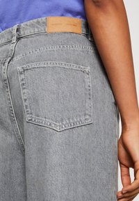 Hallitusseente denim püksid, millel on nähtav tagatasku ja tan värvi nahkstikandid vööl. Tekstuur tundub vastupidav ja klassikaline lõige.