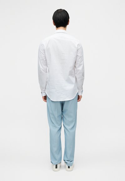 Άσπρο πουκάμισο μακρυμάνικο συνδυασμένο με ανοιχτό μπλε ριγέ παντελόνια. Τα παπούτσια είναι λευκά με πράσινες και μαύρες λεπτομέρειες. Φαίνεται από πίσω.