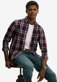 Camisa de franela a cuadros en azul marino, rojo y blanco; mangas largas remangadas; combinada con una camiseta blanca y jeans de mezclilla; sentado en un taburete de madera.
