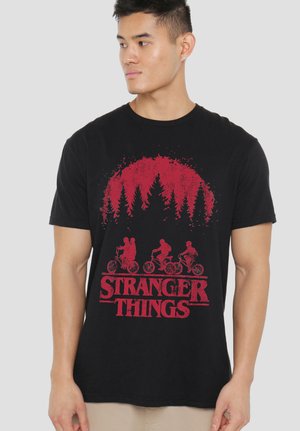 Man wearing black Stranger Things T-shirt with red graphic of three kids riding bikes beneath stylized pine trees.