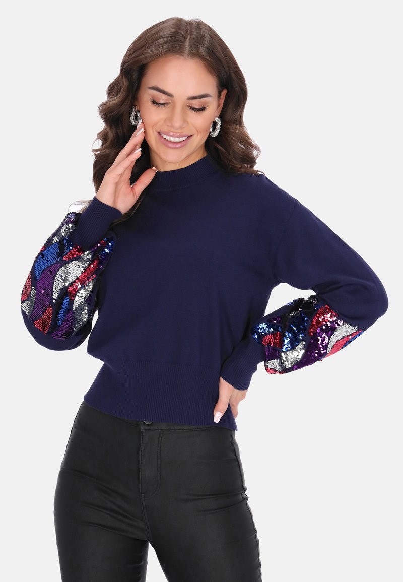 Sötétkék pulóver, amely testhez simuló fazonú, és dús ujjaiban piros, lila és ezüst színű színes flitterek díszítik. Bordázott alja és mandzsettái vannak.