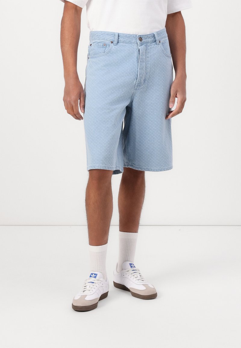 Man wearing light blue textured denim shorts, white crew socks, white sneakers with blue logo, and a white short-sleeve shirt.
