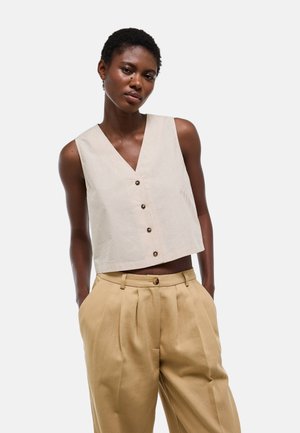 Femme portant un crop top beige sans manches boutonné et un pantalon plissé kaki taille haute, les mains dans les poches, sur fond blanc.