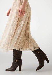 Bottes en cuir marron, hauteur genoux, avec un bout pointu, un talon block et un accent doré. Portées avec une jupe beige fluide, plissée et ornée de dentelle.