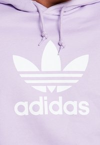 Levandulová mikina s kapucí z měkké tkaniny, s bílým logem Adidas s motivem tří listů a třemi vodorovnými pruhy pod ním.