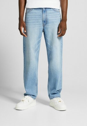 Persona vestida con jeans rectos de color azul claro y zapatillas blancas, de pie contra un fondo gris liso.