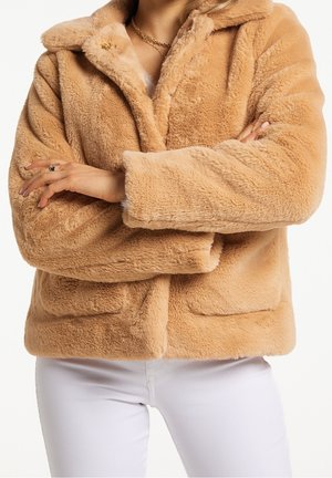 Personne portant une veste en fausse fourrure beige avec col et poches, bras croisés, associée à un pantalon blanc et un collier chaîne doré.