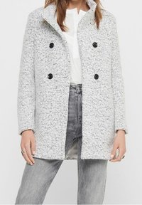 Cappotto grigio chiaro texture con bottoni neri, design doppiopetto, colletto a punte e maniche lunghe, abbinato a jeans a vita alta.