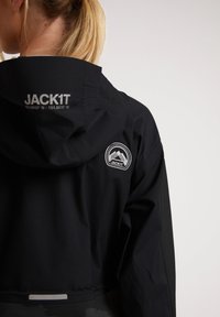 Svart vattentät jacka med huva, reflextryck och en logotyp med bergsgrafik på ryggen. Slät textur.