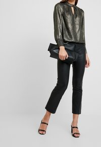 Blusa nera lucida a maniche lunghe con scollatura a chiave, abbinata a pantaloni in pelle nera aderenti e tacchi a spillo neri. Borsa a pochette in mano.
