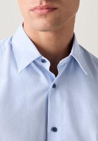 Chemise bleu clair à motifs avec un col étalé, deux boutons, et un tissu texturé. Le motif est composé de petites formes géométriques.