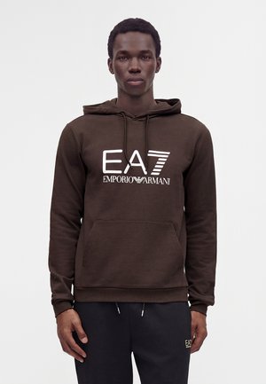 Άνδρας που φοράει σκουρό καφέ φούτερ Emporio Armani EA7 και μαύρο παντελόνι με κορδόνι, στέκεται μπροστά σε απλό ανοιχτό φόντο.