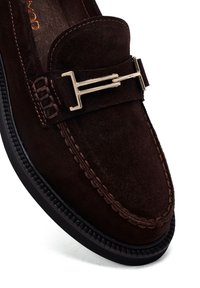 Mocassin en daim marron foncé avec une couture détaillée et une boucle métallique dorée sur le dessus, doté d'une semelle en caoutchouc noire.