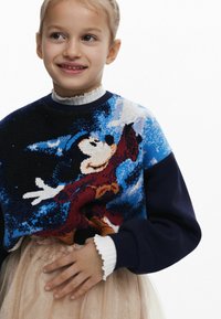 Pleteni dukserica s šarenim dizajnom Mickey Mouse-a, tamnoplave boje, s rebrastim manžetama. Nosena preko svijetle, nabirane suknje.