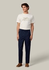 Wit T-shirt met korte mouwen en groen logo, gecombineerd met marineblauwe broek. Bruine suède schoenen maken de outfit compleet. Eenvoudig ontwerp, zonder patronen.