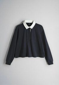 Langærmet sort poloshirt med kontrastfuld hvid krave, knapstolpe og ribstrikkede manchetter, vist mod en ensfarvet grå baggrund.