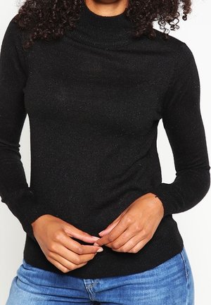 Woman wearing a black long-sleeve turtleneck sweater and blue jeans, hands clasped at waist, curly hair visible at shoulders.