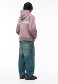 Kapuzensweatshirt in helllila mit weißem Druck auf dem Rücken, kombiniert mit lockeren blauen Jeans und braunen Turnschuhen.