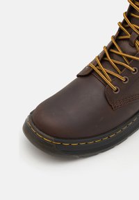 Botín de cuero marrón con cordones amarillos, puntera redondeada, acentos cosidos y suela de goma negra texturizada.