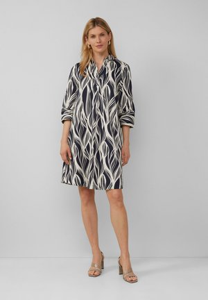 Robe marine avec un motif de feuilles crème, col polo, manches trois-quarts et coupe évasée. Associée à des sandales à talons beiges.