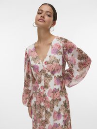 Vrouw in een doorzichtig jurk met lange mouwen, versierd met een bloemenprint in roze, paars en bruin, met een V-hals en knopen aan de voorkant.