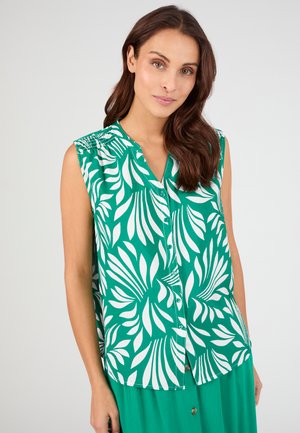 Femme portant un chemisier vert sans manches avec des motifs de feuilles blanches et un col en V, associé à une jupe verte assortie, sur un fond uni.