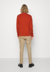 Personne aux cheveux blonds mi-longs portant un pull couleur rouille, un pantalon beige et des chaussures noires, debout contre un mur blanc.