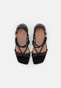 Schwarze High Heels Sandalen mit quadratischem Zehenbereich und kreuzenden elastischen Riemen. Weiche Innenauskleidung in Beige, mit goldenem Markenlogo.