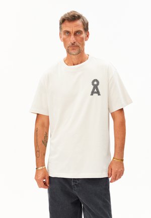 Weißes Baumwoll-T-Shirt mit einem strukturierten grauen Banddesign auf der linken Brust. Kurze Ärmel und eine lockere Passform. Kombiniert mit dunklen Jeans.