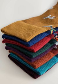 Seks par foldede sokker i forskellige farver: sennep, marineblå, rød, grøn, lilla, teal og orange. Strikket tekstur med mønstrede detaljer.
