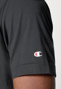 Schwarzes Baumwoll-T-Shirt mit kurzen Ärmeln und einem rot-weißen Logo am linken Arm. Glatte Textur und lässiges Design.