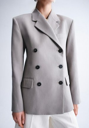 Mujer con un blazer gris claro de doble botonadura con botones negros y pantalones blancos, posando frente a un fondo liso.