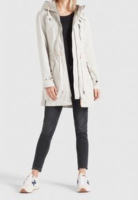 Giacca traspirante di un grigio chiaro con cappuccio, vita regolabile con coulisse e tasche frontali con zip, abbinata a jeans neri e scarpe da ginnastica chiare.