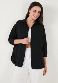 Black button-up shirt with a relaxed fit, featuring a textured fabric, collar, and rolled sleeves, paired with white pants.