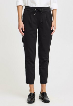 Schwarze Jogginghose mit verstellbarem Bund, zwei Fronttaschen, seitlichen Streifen und schmalem Schnitt. Getragen mit schwarzen Lackloafers.