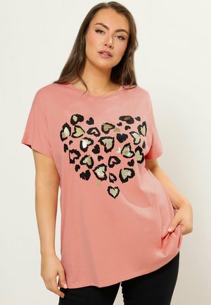 Kvinde iført en løstsiddende lyserød T-shirt med sorte og gyldne leopardprint-hjerter, der danner et stort hjerte foran.